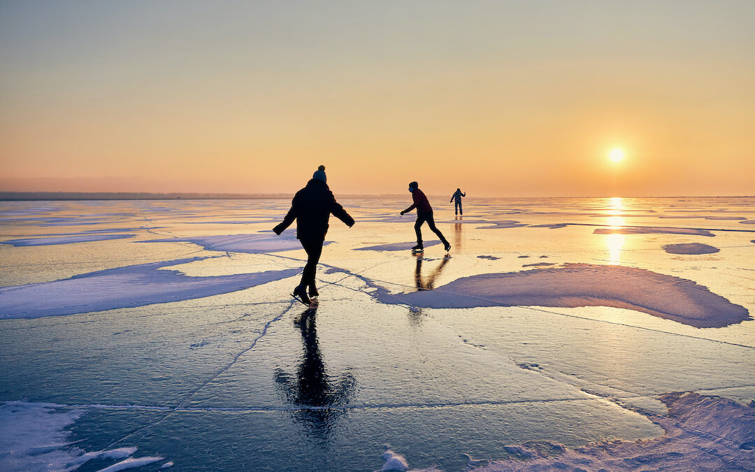 Image is of ice skaters on a frozen lake, skating toward he sunrise.