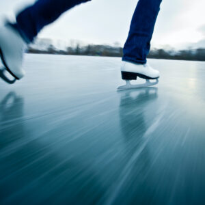 Closeup of the white ice skates of a young woman ice skating outdoors on a the surface of a frozen pond, illustrating an essay about moving through life with as much control as we can.